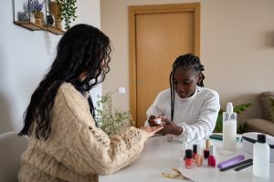 DIY Nail Care. Young woman painting nails of her friend, sitting at table with nail polishes, files and other manicure tools. Is DIY Nail Care Hurting Your Salon Business?