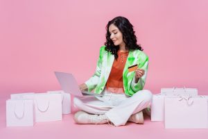 pleased woman in blazer using laptop while holding credit card near shopping bags on pink. Top 3 Online Nail Supply Stores Canadians Trust in 2025.
