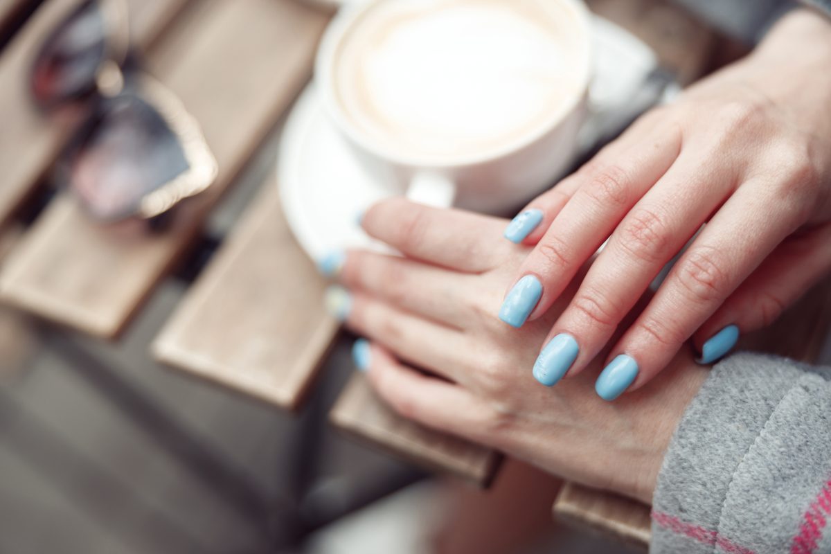 Top view on female hands with blue manicure and cup of cappuccino at table outdoors. Close up.