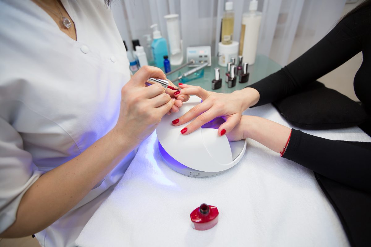 Close-up of the hand of a manicurist who applies red gel Polish on the nails of a young girl. Girl dries gel nail Polish in UV lamp.