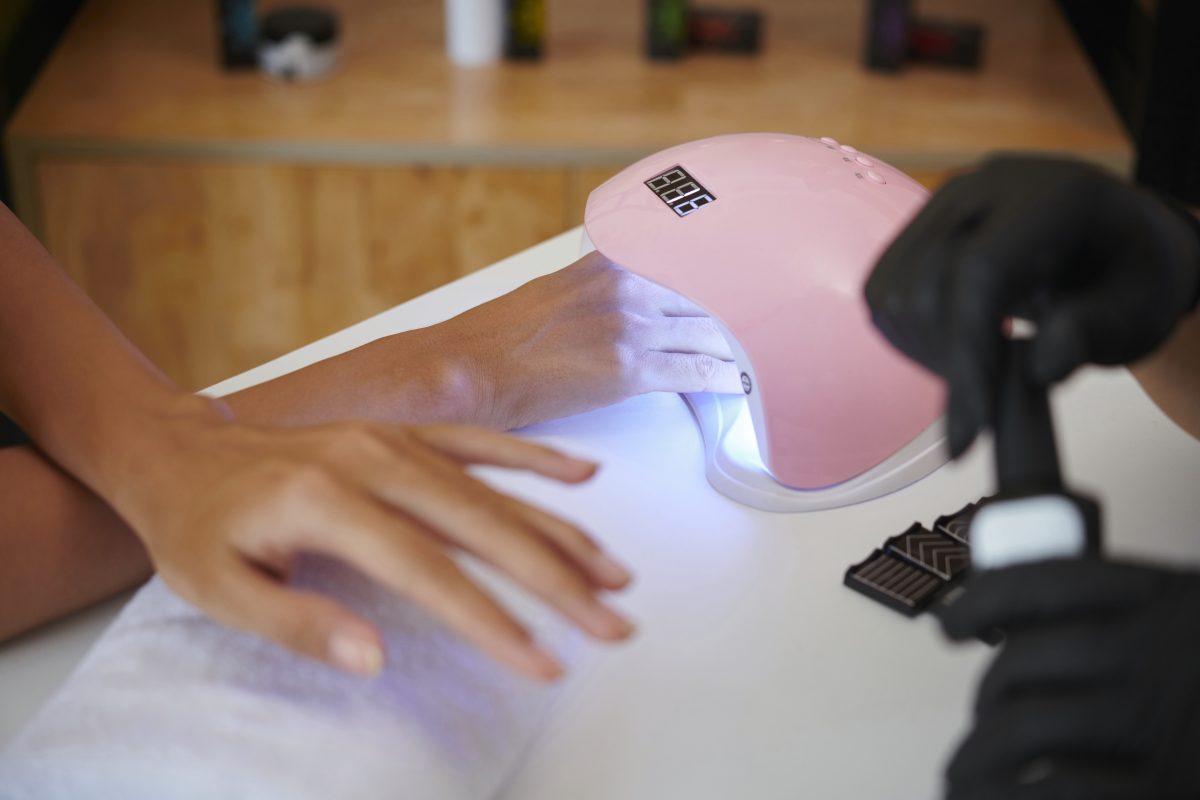 Hands under UV light device being used for nail curing in professional salon setting with various tools on table capturing moment of beauty treatment