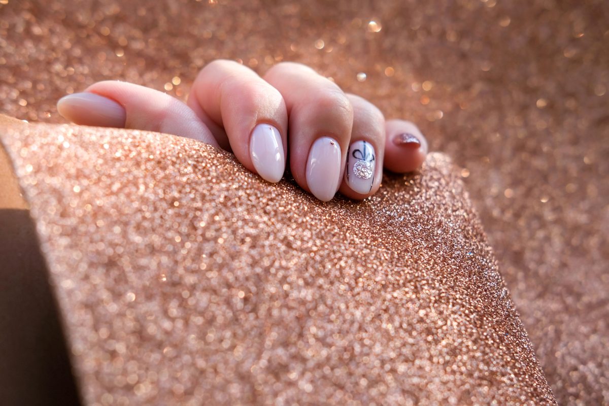 Close-up of a manicured hand with a pink and silver glitter gel design featuring holiday-themed nail art, resting on a textured purple knit background with silver star confetti.