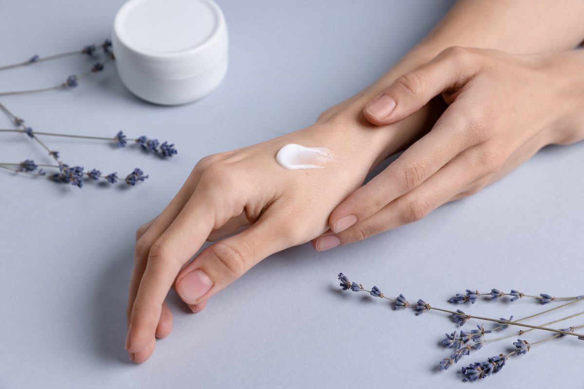 Woman with hand cream and lavender flowers on grey background, closeup